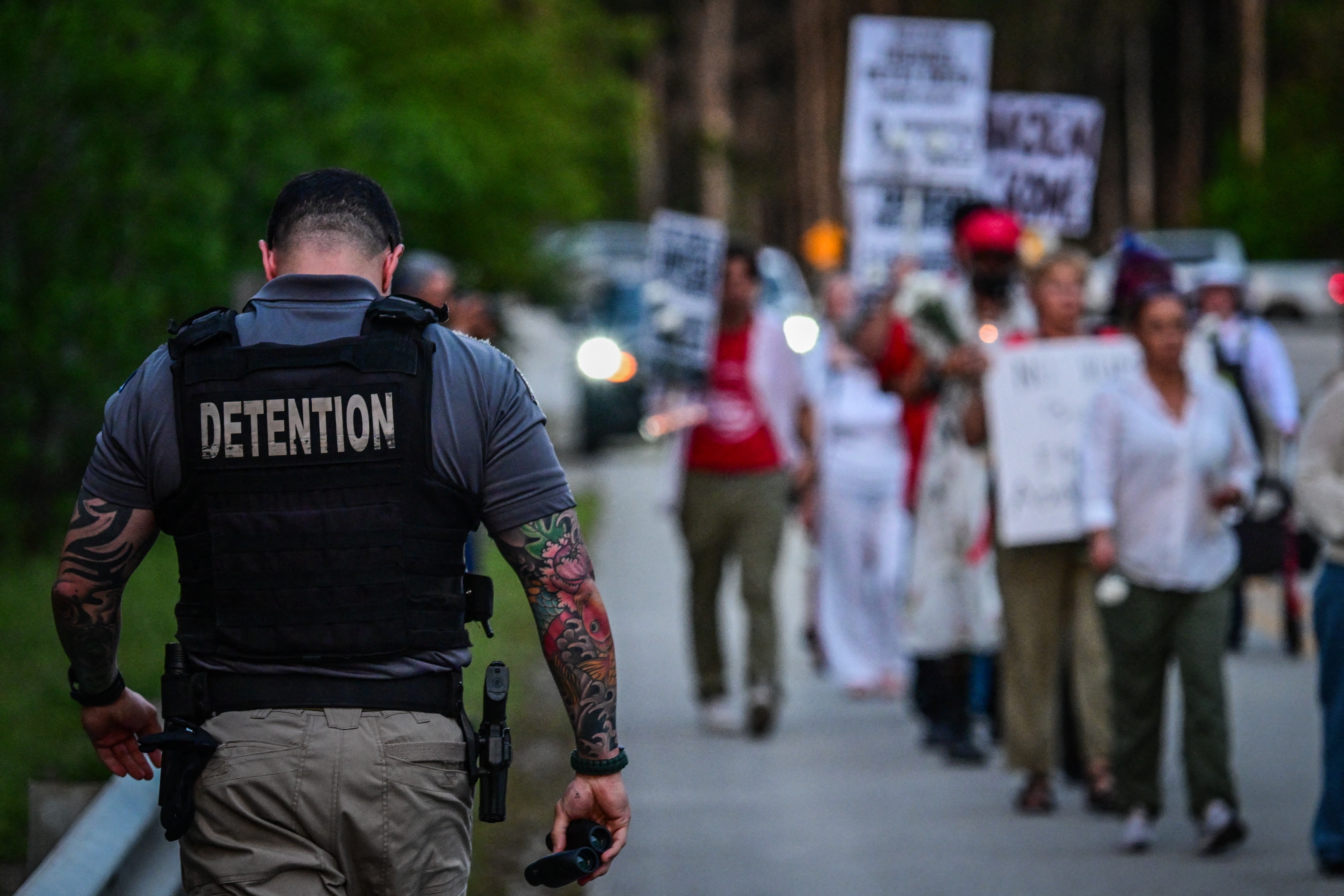 A corrections officer walks beside people holding candles, signs, and flowers during a vigil outside the Krome Detention Center in Miami in May 2025, protesting U.S. Immigration and Customs Enforcement custody and mass deportations.